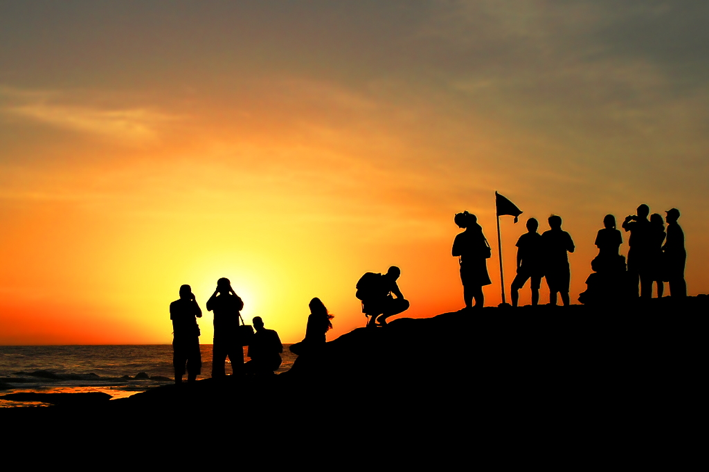 Silhouettes of people at the top of a hill, gathered by a flag and taking photos of an orange sunset