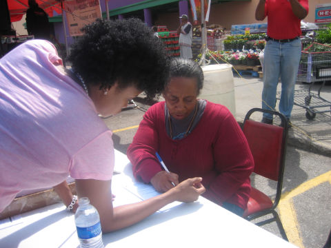 A young Black woman leans over a table, helping another older Black woman fill in a form