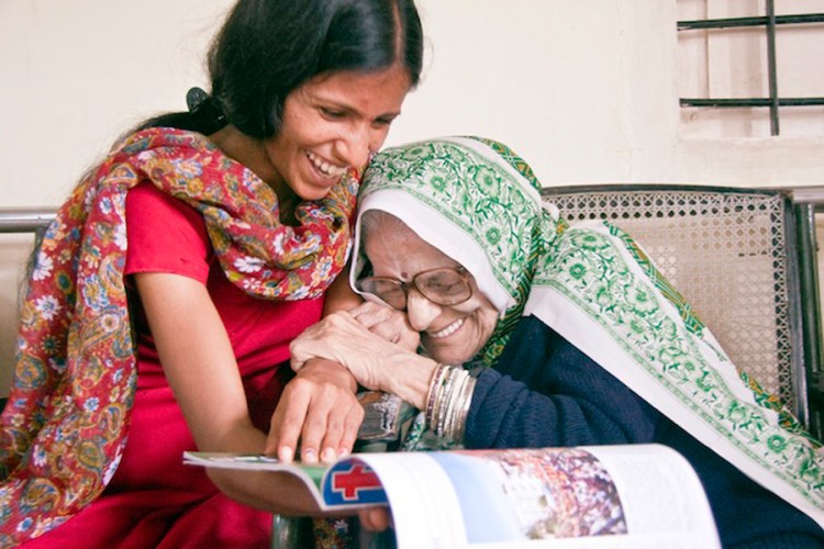 Two Indian women laugh and grip one another, one younger with long black hair is holding a magazine, while the older woman who wears a colourful green scarf is hunched over in joy