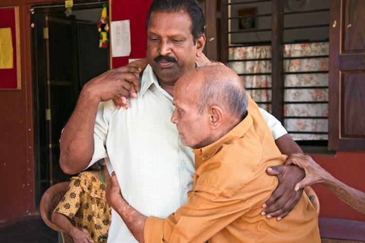 Two Indian men, one middle aged with short black hair supports an elderly man who is balding, and hugging the other man