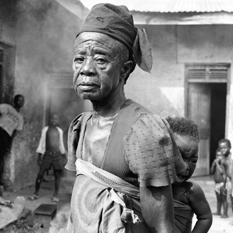 Black and white photo of an elderly Black woman has a crying baby strapped to her back
