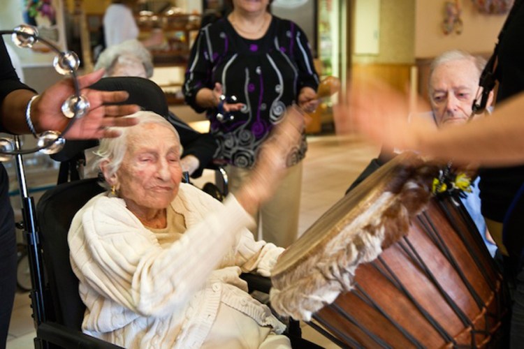 An elder white woman claps as muscians play a drum and tambourines, while a man watches