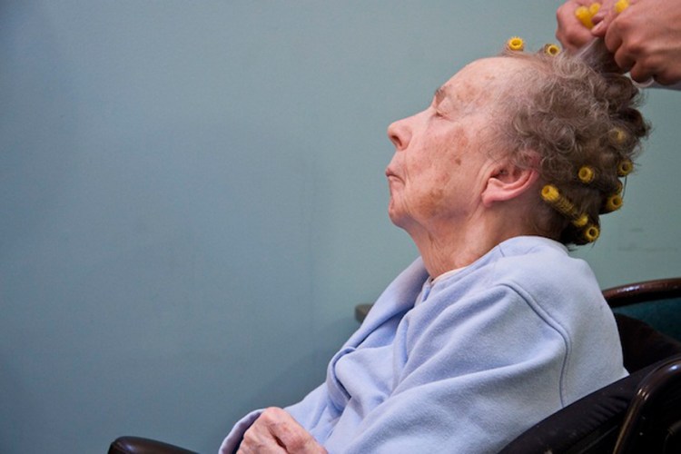An elderly white woman is getting her hair put into rollers, while her head is back and her eyes are semi-closed