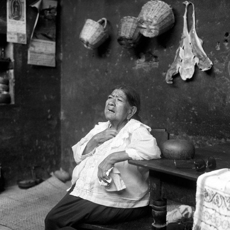 An elderly Peruvian woman is sitting with her hand on her heart, as she looks up with her mouth open