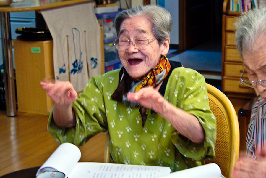 An elderly Japanese woman is seated, smiling, moving her hands, and singing in a colourful room