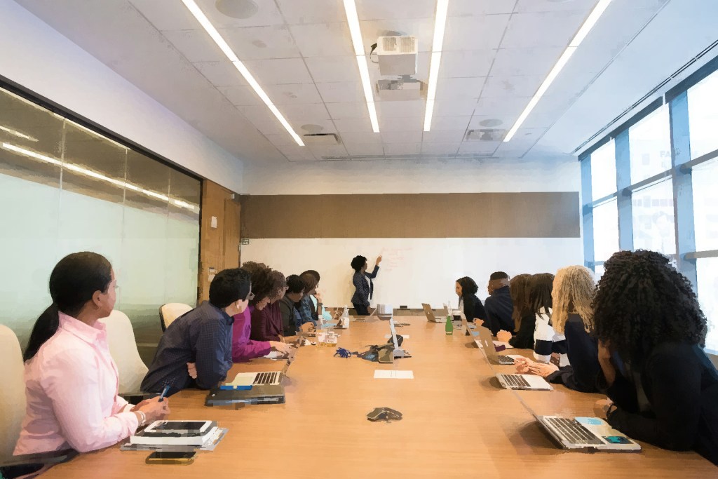 A Black woman is presenting to a large diverse group sitting at a board table. She points to a white board 