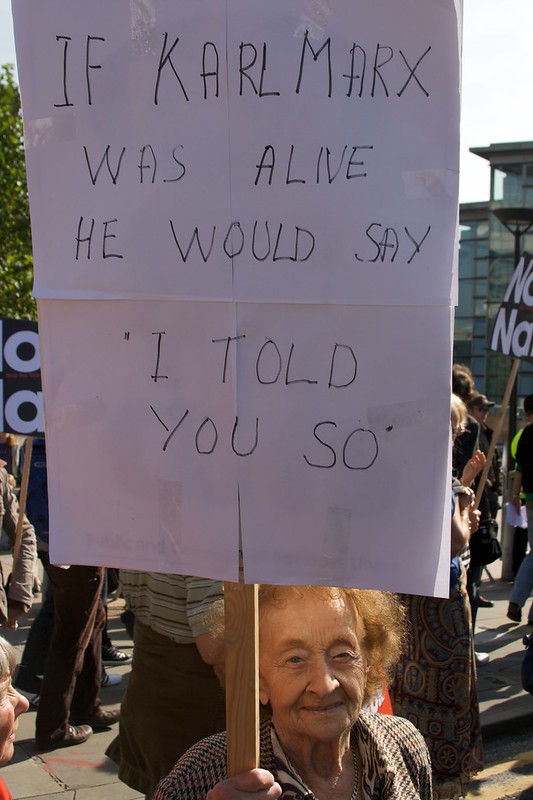 An elderly woman holds a sign and smiles. It reads: If Karl Marx was alive he would say, I told you so