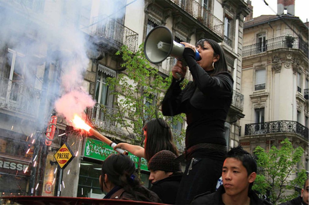 Woman speaking into megaphone at a protest with young people underneath, one is holding a light