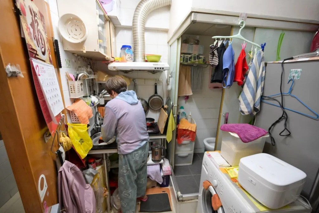 A person washes dishes in a tiny apartment, next to the washing machine and toilet