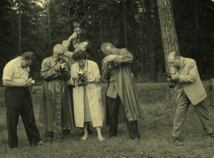Zofia Rydet is a young white woman with short dark curly hair. She stands in the centre wearing a white full-length coat. She is surrounded by five male photographers. They are all looking at their cameras, standing in the woods