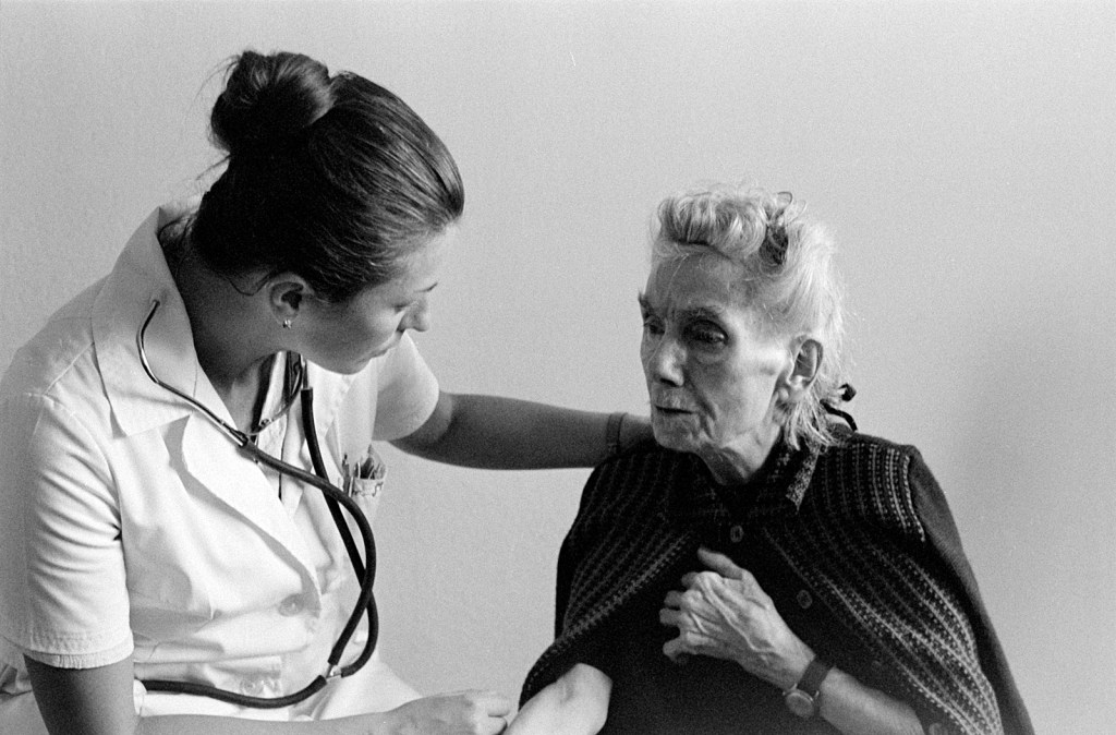 Black and white photo of two white women. One is a nurse looking concerned, with her arm around a frail patient