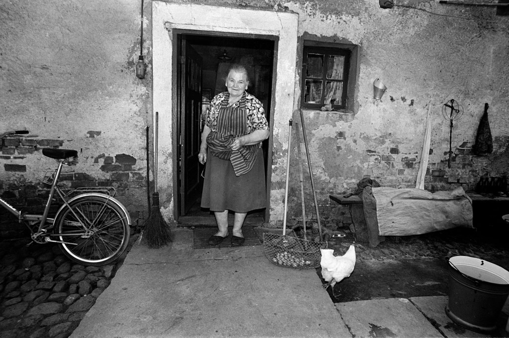 Black and white photo of an older white woman who is smiling at her doorway. A chicken is walking on the right and a bike is on the left