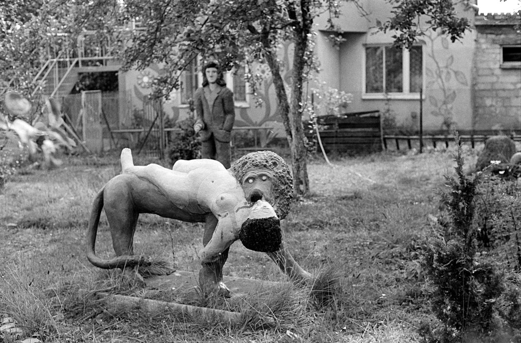 Black and white photo of a statue of a person being eaten by a lion, with another statue of a man in the middle of a large garden