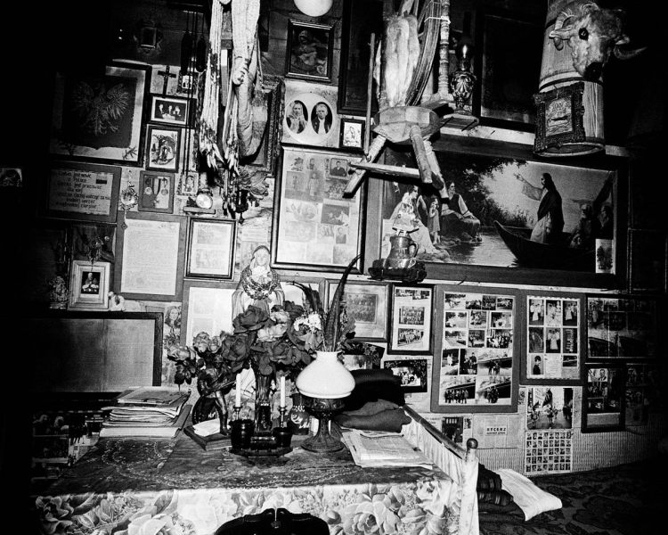 Black and white photo of a wall crowded with photographs and handing statues, as well as a table decorated with a saint figurine, flowers, and papers