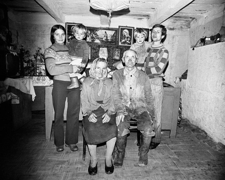 Black and white photo of a family. An elderly man and woman sit, with a younger woman and man standing holding twin boys. Behind them are various biblical framed pictures and figurines