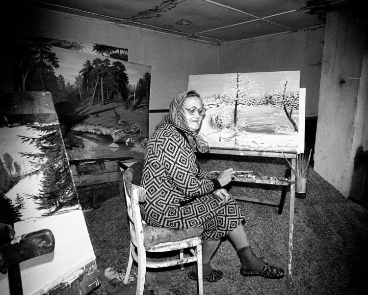 Black and white photo of a woman in a scarf and pattered dress paining a snow scene. She is surrounded by paintings in a water-damaged room, with stains on the walls and roof