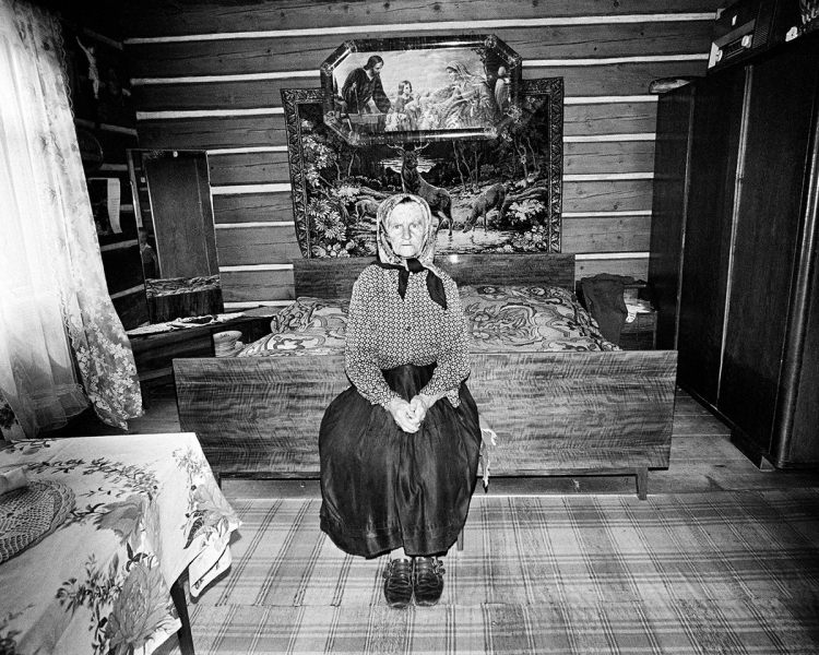 Black and white photo of an elderly woman scowling in a bedroom. She wears a scarf around her head, a diamond-pattern blouse and a long dark skirt. She is in frong of a large wooden bed and two large paintings, one showing a deer by a creek, and the other of a religious scene, likely depicting Jesus, Mary and Joseph