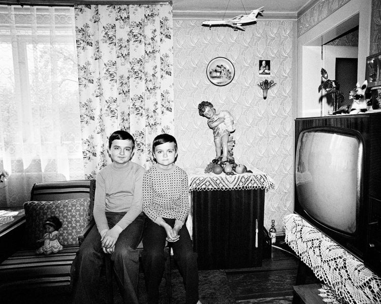 Black and white photo of twin boys with small smiles. They are sitting on chairs surrounded by a large CRT TV from the 1980s. There are various figurines and a white model plane hangs from the ceiling