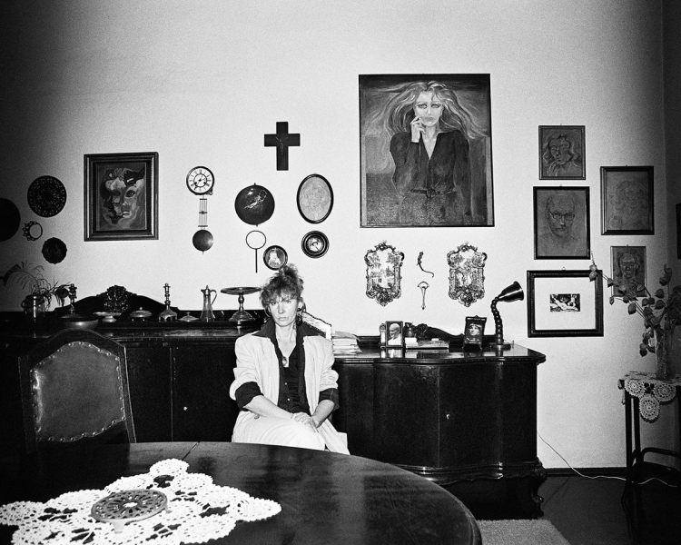 Black and white photo of a middle-aged woman who is not smiling. She sits in a large living room with dark wooden furniture and various expressionist paintings in the background, as well as a large crucifix and photographs