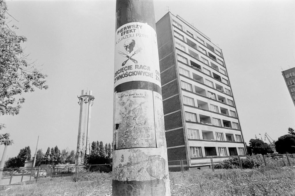 Black and white photo of a pole with socialist posters. A multi-storey building is the background
