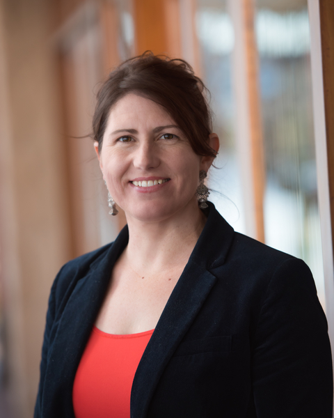 Dr Samantha Crompvoets is a white woman with light brown hair tied back. She wears a navy blazer with a red top and earrings. She is smiling.