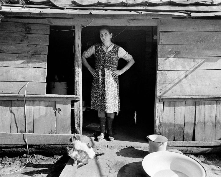 Black and white photo of a middle-aged woman with her hands on her hips. She stands under a wooden awning, with two cats by her feet and large water containers