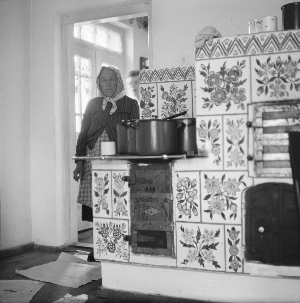 Black and white photo of a woman waring a scarf standing at the doorway of a kitchen, with a man out of focus behind her. The stove is made of beautiful ceramic tiles with painted flowers