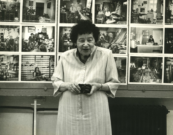 Zofia Rydet is an elderly white woman with short dark curly hair, wearing a long dress with vertical stripes. She is holding a small electronic camera. She stands in front an exhibition of her work