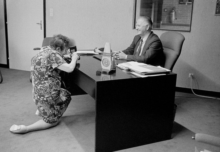 Zofia Rydet is an elderly white woman with short curly hair. She is kneeling and smiling as she photographs a white older man who is smiling behind his office desk