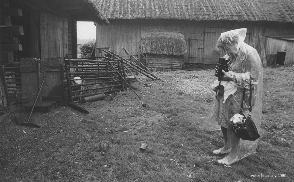 Zofia Rydet is an elderly white woman wearing a full-length raincot and a knee-length dress with white shoes. She is taking a photo outdoors, next to two large cabins
