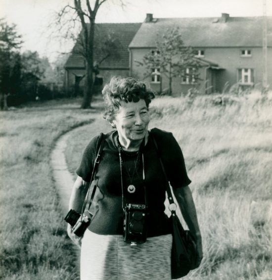 Zofia Rydet is an older white woman with short light curly hair. She is smiling as she walks in a field. She has two cameras around her neck, and a bag around her shoulder. A large rural house and tree is in the background