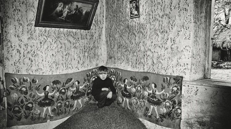Black and white photo of a young boy crouching in the corner of a room with ornate wallpaper. The scene portrays art of women dancing surrounded by flowers. Two biblical paitings hang above. To the right we see outdoors, to a hut with a ladder reaching over the roof