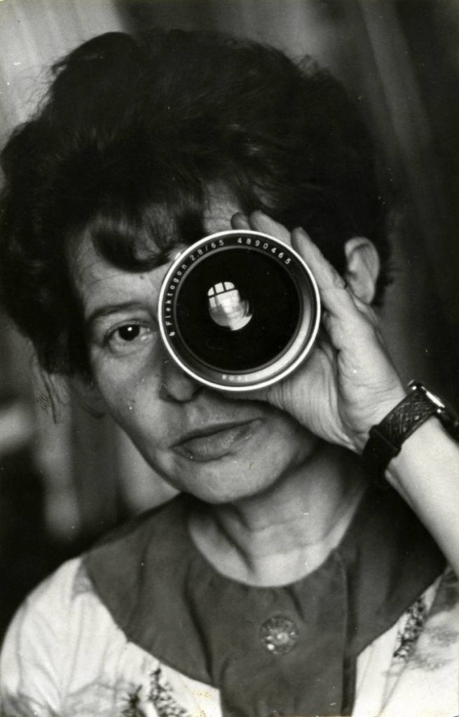 Zofia Rydet is a middle-aged white woman with dark wavy hair done up in a beehive. She looks through a camera lens with a determined expression