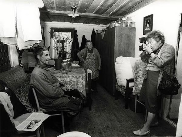 Zofia Rydet is an older white woman with short light curly hair. She stands against a wall taking the photo of a man who is seated. Another older woman watches from the back of a small, crowded living room