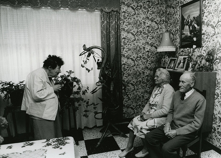 Zofia Rydet is an elderly white woman with short dark curly hair. She has a hunched back. She is speaking with an elderly couple who are seated having their photograph taken. They are in a room with ornate flower-patterened wallpaper, and a folkloric painting of a man and a woman in the background, and various photos on a mantel