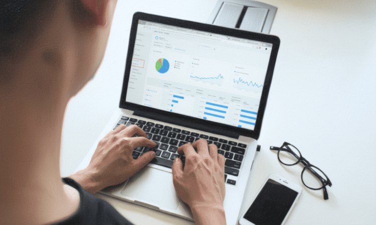 The back of a white man's head. He is typing on a laptop showing graphs, with reading glasses and a mobile phone beside him