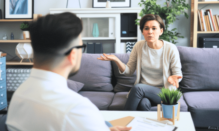 A white woman sits on a coach with her arms out as if asking a question. A white man has his back to us, holding a clipboard