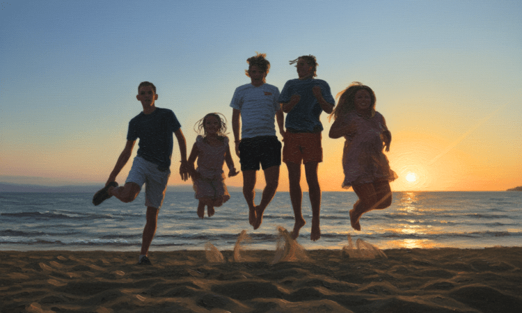 Five youth jump in the air on a beach, their feet kicking up sand. The sun is setting in the background