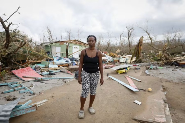 A young Black woman stands in front of broken wood and a fallen house