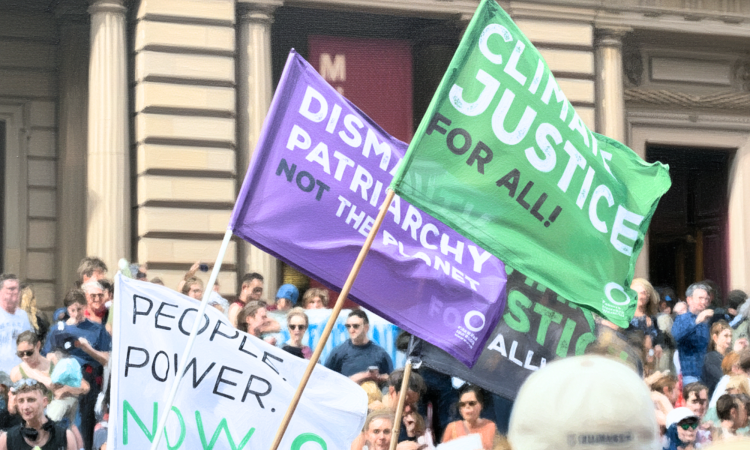 Protesters are crowded in the background in front of the a large building. Close of banners saying 'Climate justice for all!,' 'dismantle the patriarchy not hte planet,' and 'people power now.'