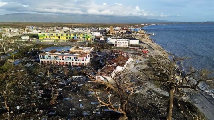 Aerial view of a ruined hotel without a roof is the only building left near the coast, with threes and houses broken around it