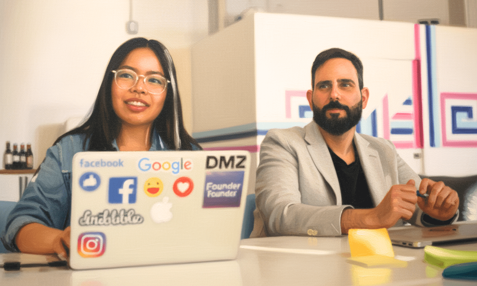 An Asian woman and white man sit at a table looking to the side, with their laptops in front of them