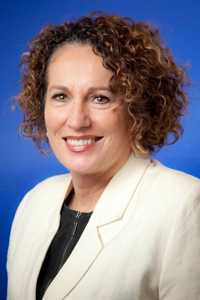 Dr Helen Szoke is a white woman with short, auburn, curly hair. She wears a white jacket and is smiling against a blue background