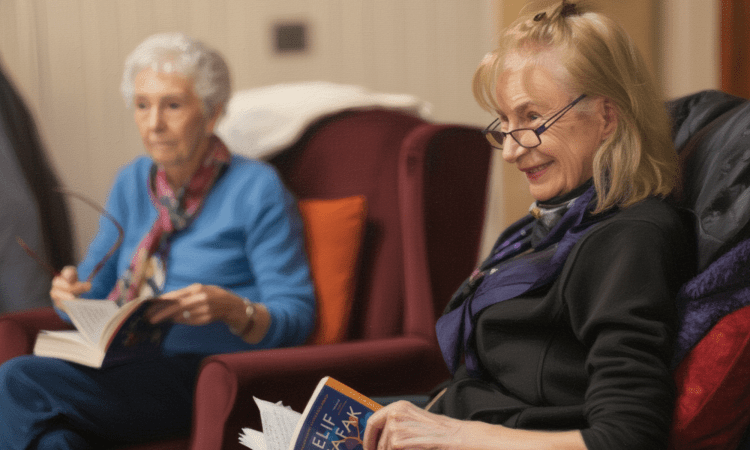 Two white elderly women sit on chairs. In the foreground, she has blonde and grey hair, and wears glasses, she is smiling holding a book by Elif Shafak