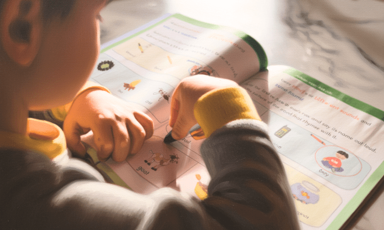 A white child draws in a workbook, circuling a picture and word for 'goat'