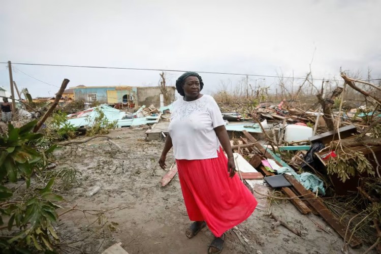 A Black woman looks to the side with broken wood and shattered houses in the background