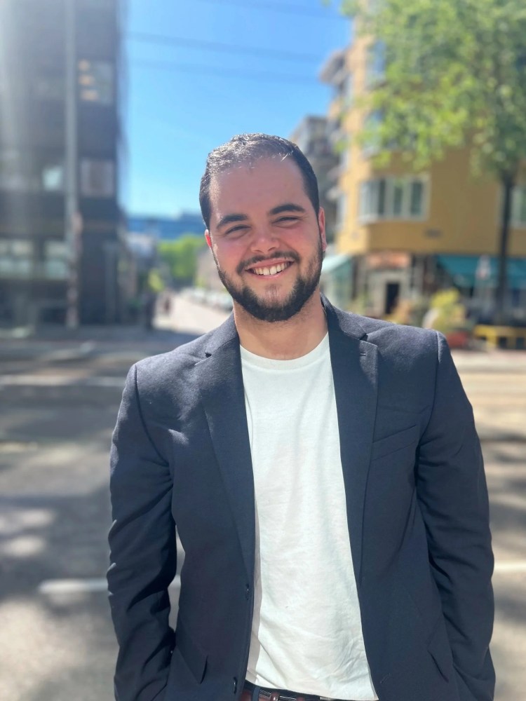 Parker Lemal-Brown is a white man with black hair and a beard, wearing a blue jacket and white t-shirt, smiling in the street