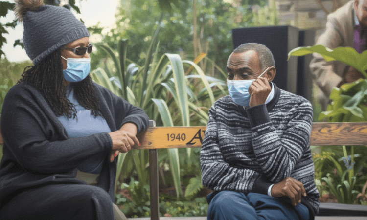 A Black man and woman sit outdoors on a park bench. Both wear face masks. They are talkng and smiling
