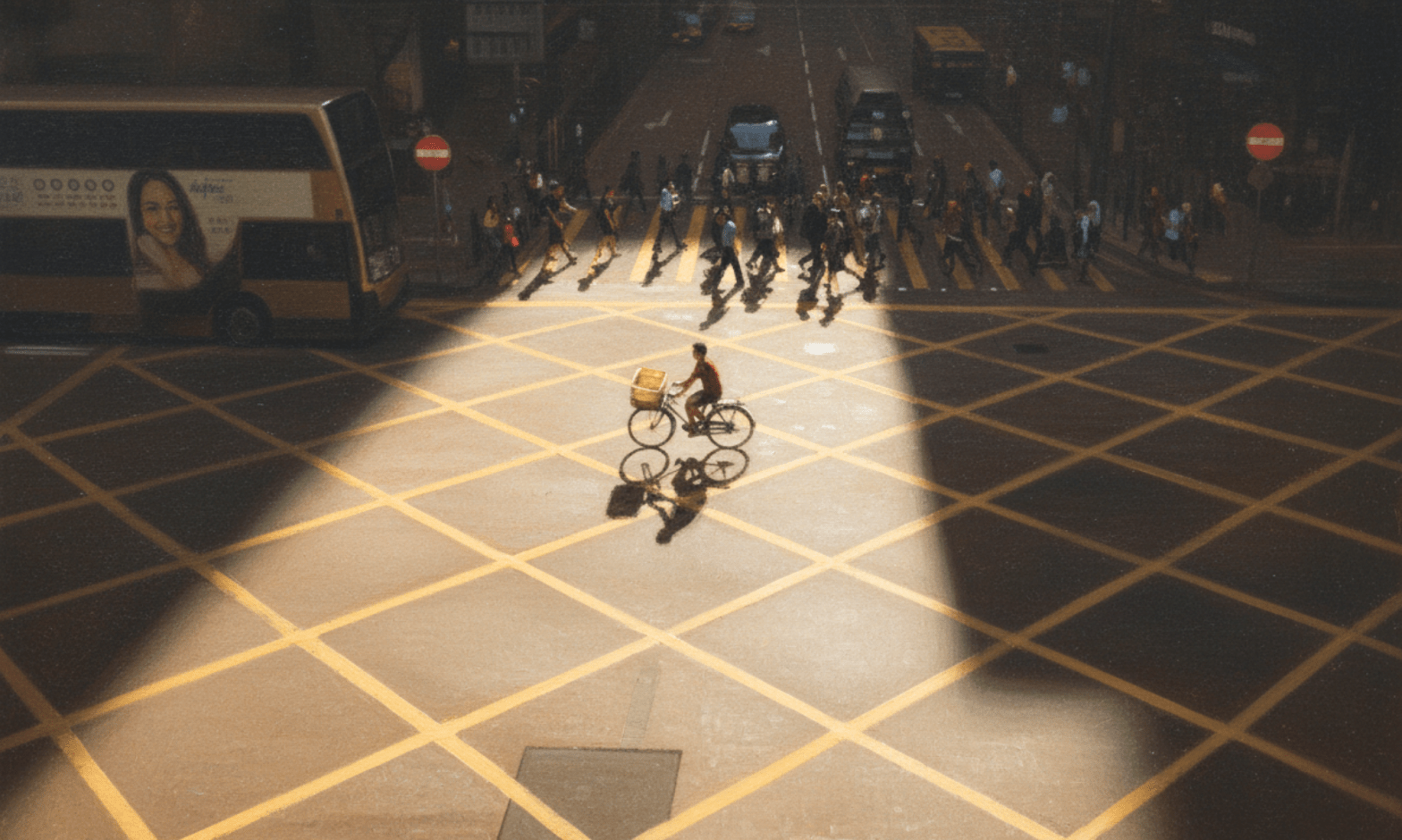 A lone man rides his bike across an empty road, with pedestrians and cars in shadow in the background