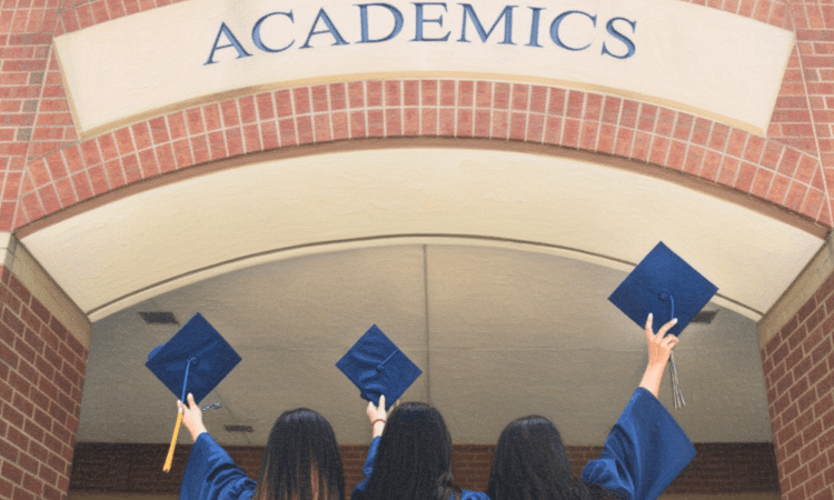 Three women with their backs to us hold us their blue graduation caps in front of a building that says, 'ACADEMICS'
